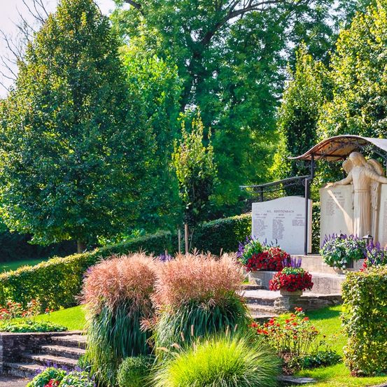 Eine Statue mit zwei Figuren steht unter einem Baldachin in einem gepflegten Garten mit blühenden Pflanzen und ordentlich geschnittenen Hecken. Bäume und Büsche umgeben den Bereich und schaffen eine ruhige und friedliche Atmosphäre.