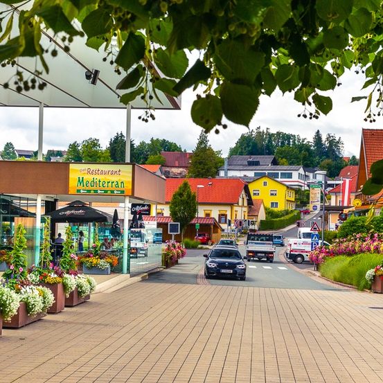 Eine belebte Straße mit bunten Häusern und viel Grün. Ein Restaurant mit mediterraner Küche befindet sich vorne, mit geparkten Autos und Blumen an der Seite.
