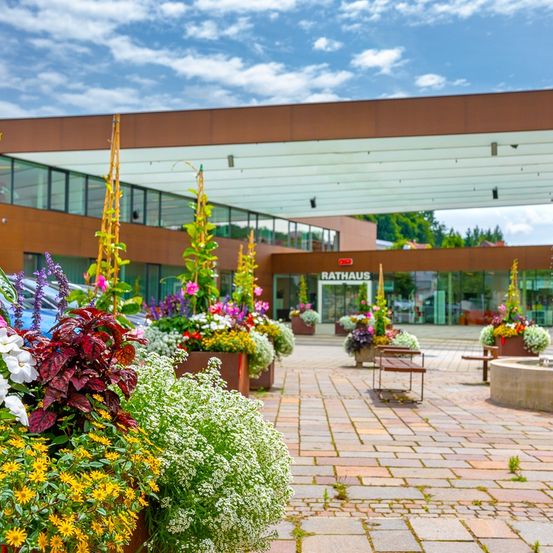 Das Rathaus zeigt ein modernes Gebäude mit großen Glasfenstern. Ein blumenreicher Vorhof führt zum Eingang.