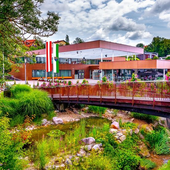 Ein modernes Gebäude mit Fahnen steht in der Nähe einer Brücke über einem Bach, umgeben von üppigem Grün und bunten Blumen.