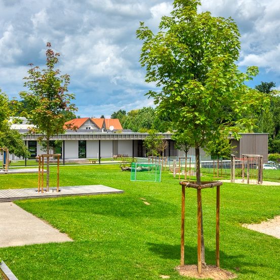 Ein grasbewachsenes Feld mit mehreren jungen Bäumen, einem großen weißen Gebäude mit orangem Dach und verschiedenen Spielgeräten, unter einem teilweise bewölkten Himmel.