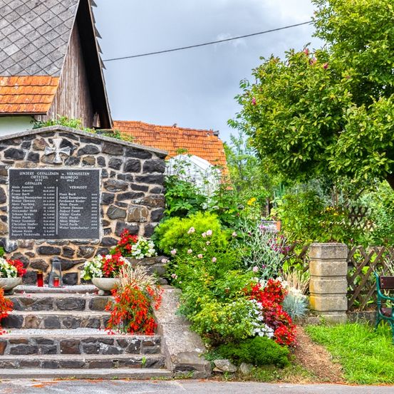 Ein Gedenkstein mit einem Kreuz und Namen gefallener Soldaten steht auf einer Steinplattform. Blumen und Pflanzen umgeben das Denkmal. Eine Bank ist am Rande des Grases platziert.