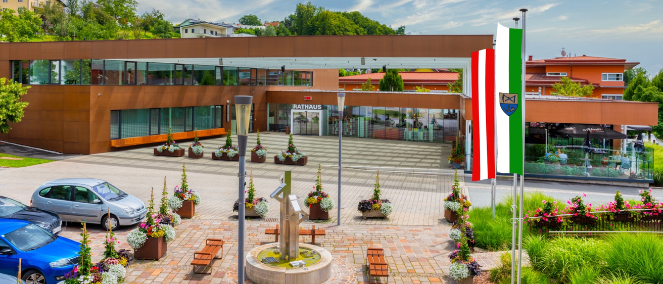 Ein Gebäude mit Glaswänden steht im Zentrum der Stadt. Ein Auto ist vor dem Gebäude geparkt. Eine Brunnenanlage befindet sich in der Mitte des Gehwegs. Auf der rechten Seite stehen Fahnen auf Stangen.