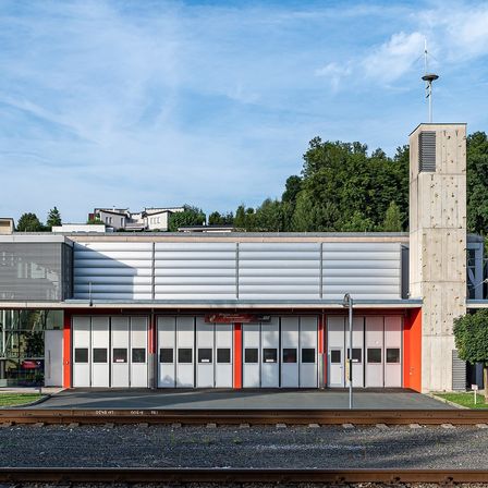 Ein modernes Feuerwehrhaus mit Betonaußenwänden, roten und weißen Akzenten und mehreren großen Garagentoren an einem sonnigen Tag.