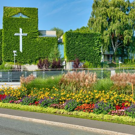 Ein Gebäude, das mit Efeu bedeckt ist, mit einem weißen Kreuz auf der Vorderseite, neben einem Garten mit bunten Blumen und Büschen. Bäume und ein Zaun sind im Hintergrund.