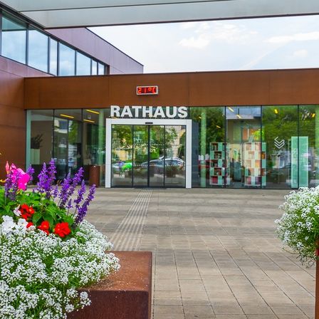 Der Eingang zum Rathausgebäude ist mit einer Glastür versehen, umgeben von Blumen, mit einer Temperaturanzeige darüber. Das Äußere hat Glasfenster und ist unter einem klaren Himmel.