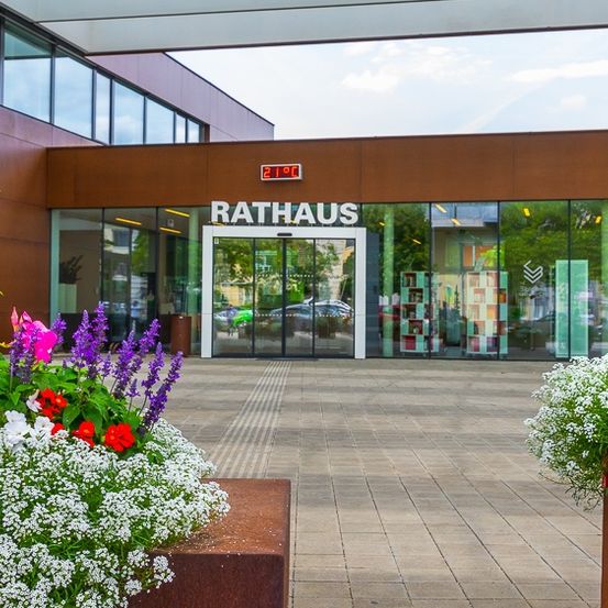Der Eingang zum Rathausgebäude ist mit einer Glastür versehen, umgeben von Blumen, mit einer Temperaturanzeige darüber. Das Äußere hat Glasfenster und ist unter einem klaren Himmel.