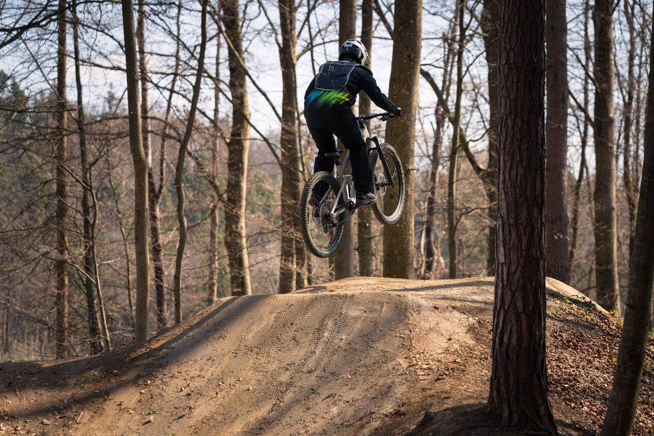Ein Radfahrer mit Helm und Rucksack ist über einem kleinen Hügel im Wald in der Luft.