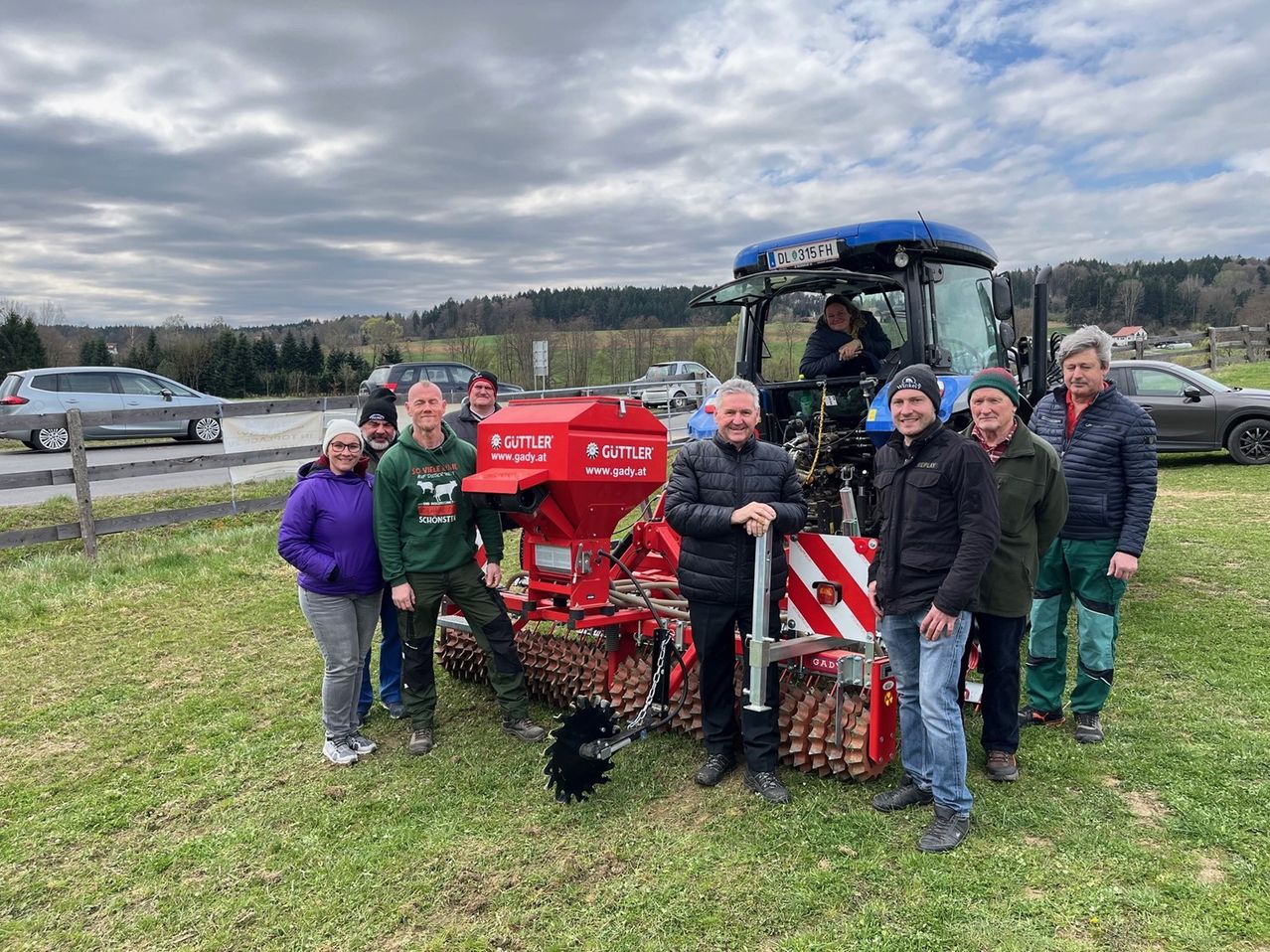 Eine Gruppe von Menschen, sowohl Männer als auch Frauen, steht vor einem roten Traktor. Sie alle lächeln und scheinen für ein Foto zu posieren. Der Traktor hat einen roten Kasten, der daran befestigt ist.