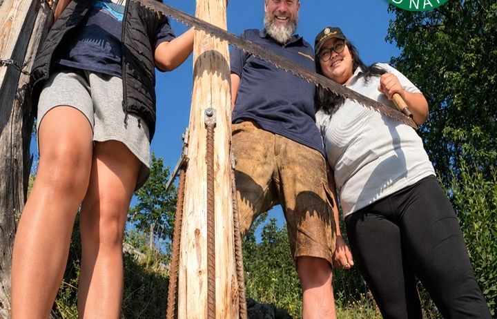 Drei Personen posieren mit einer großen Säge an einem Holzpfosten in einer Waldumgebung. Der Mann in der Mitte trägt einen Hut und ein blaues Hemd.