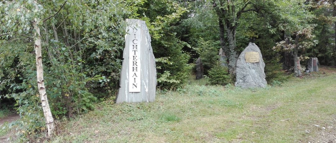 Ein großer Steinmonument mit der Aufschrift 'Dichterhain' steht in einem Grasbereich mit mehreren Bäumen. Ein weiteres Steinmonument ist im Hintergrund zu sehen.