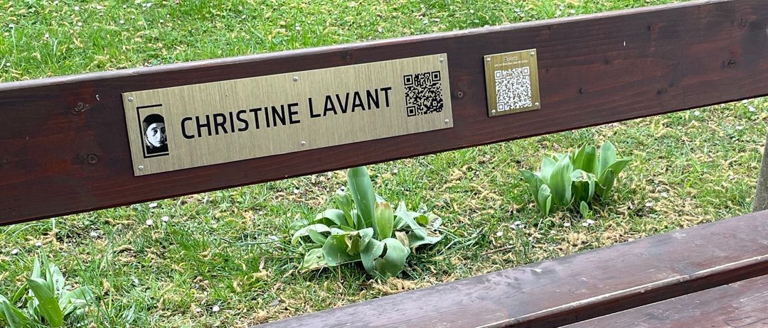 Eine Holzbank in einem Park mit einer Plakette, die Christine Lavant liest. Es gibt einen QR-Code und ein Bild einer Person auf der Plakette.