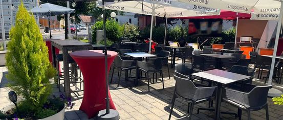 An outdoor dining area with tables and chairs under umbrellas, featuring a red table with a potted plant nearby. The area is surrounded by greenery and has a clear view of the sky.