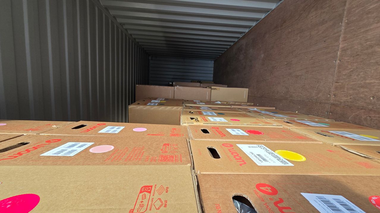 Ein Lagercontainer gefüllt mit zahlreichen Kartons, jeweils mit Etiketten und Barcodes, einige mit roten Logos und Aufklebern. Die Kartons sind in Reihen gestapelt.