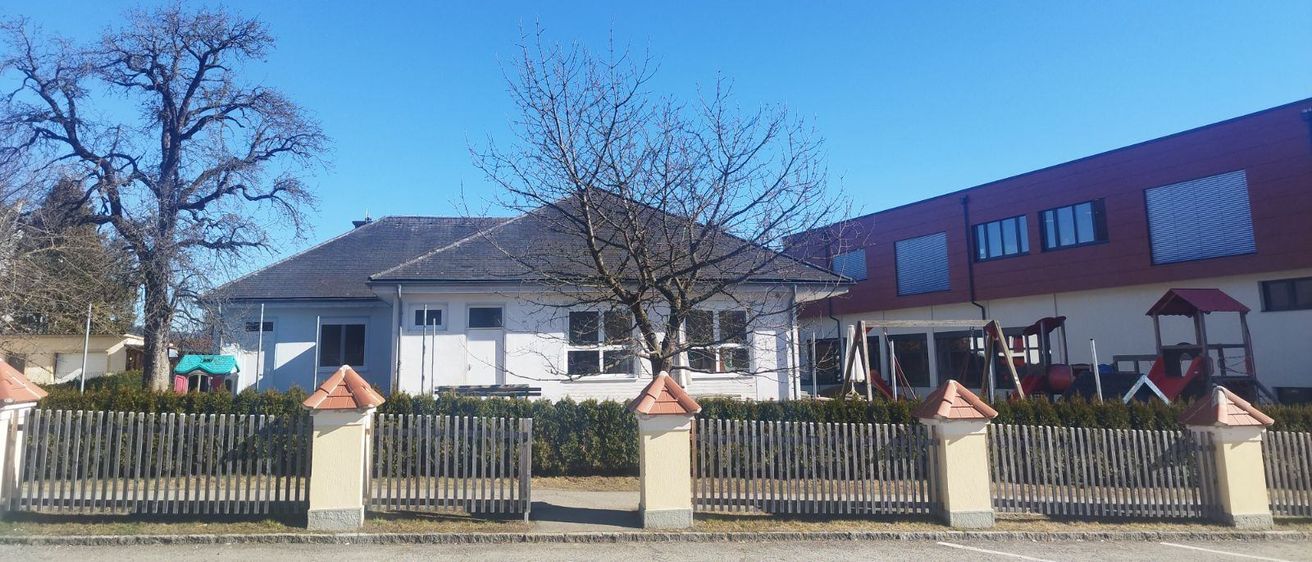 Ein weißes Haus mit grauem Dach und einem kleinen Baum davor. Das Haus ist von einer Hecke und einem Zaun umgeben. Hinter dem Haus befindet sich ein weiteres Gebäude mit Glasfenstern.