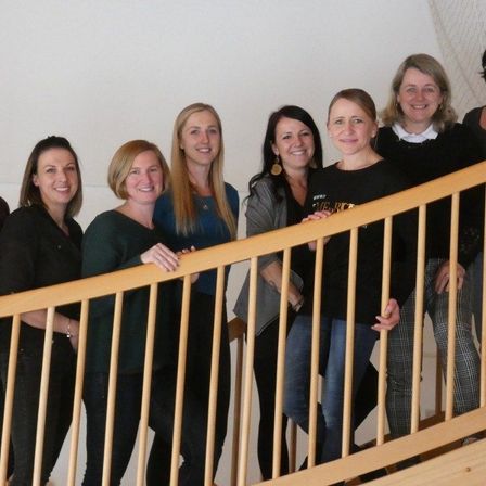 Sieben Frauen stehen auf einer Treppe und lächeln. Sie tragen lässige bis formelle Kleidung, was auf eine professionelle Umgebung hindeutet. Die Frau in der Mitte trägt ein schwarzes T-Shirt mit Text.