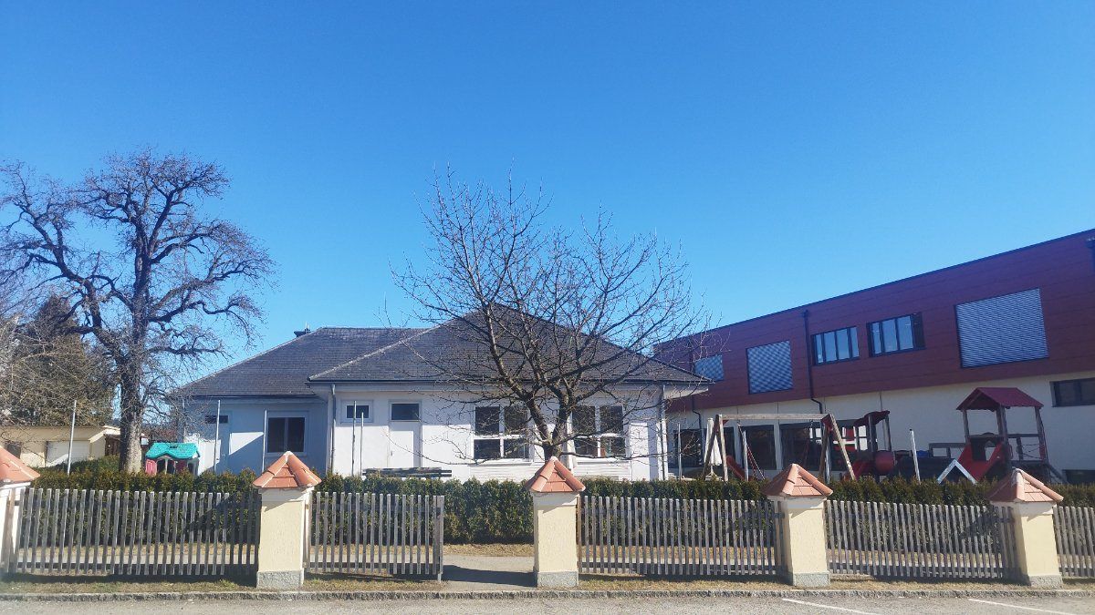 Ein weißes Haus mit grauem Dach steht vor einem Spielplatz mit einer Rutsche. Das Haus ist von einem Zaun umgeben, mit einem Tor und Pfosten. Bäume und Büsche stehen vor dem Haus.