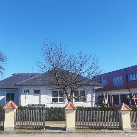 Ein weißes Haus mit grauem Dach steht vor einem Spielplatz mit einer Rutsche. Das Haus ist von einem Zaun umgeben, mit einem Tor und Pfosten. Bäume und Büsche stehen vor dem Haus.