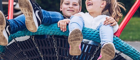 Zwei Kinder spielen auf einer Schaukel, ein Junge und ein Mädchen, beide lächeln. Der Junge trägt Turnschuhe und das Mädchen ein weißes Hemd. Die Schaukel ist blau und schwarz. Das Bild hat einen Wasserzeichen mit dem Text 'TPG'.