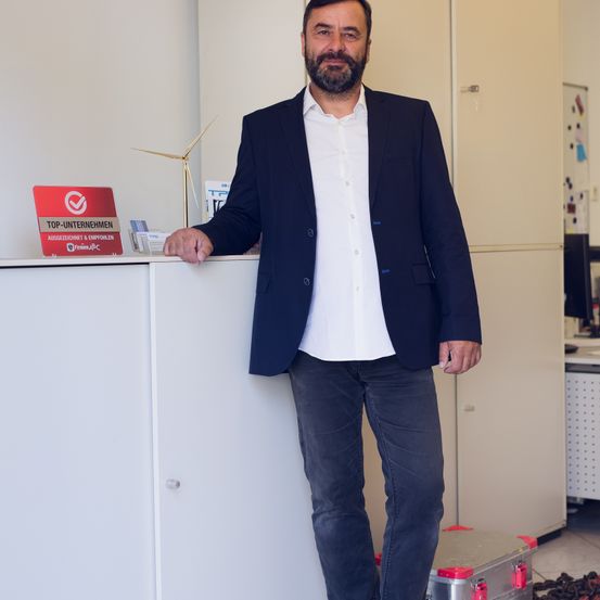 Ein Mann in einem Anzug steht an einem Tresen in einem Büro. Er hat einen Aktenkoffer neben sich und ein Schild hinter sich.