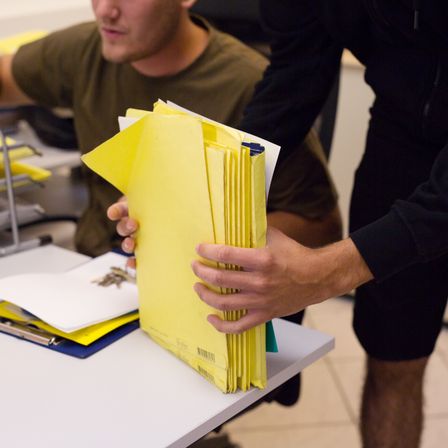 Eine Person in Schwarz hält einen Stapel gelber Umschläge, während eine andere Person in Braun hinter ihr sitzt. Ein Schreibtisch mit Papieren und Schlüsseln ist im Hintergrund zu sehen.