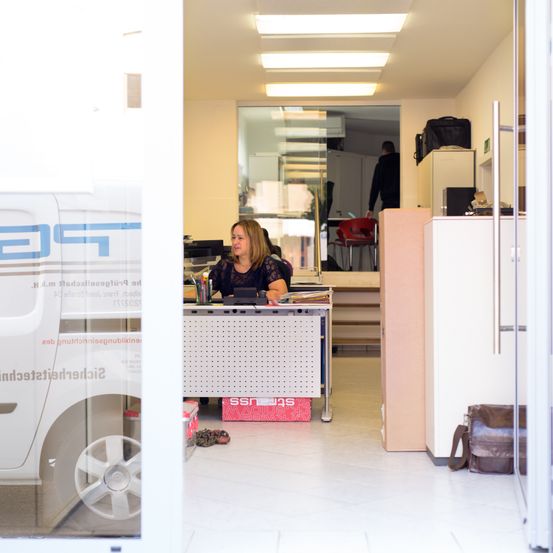Eine Frau sitzt an einem Schreibtisch in einem Büro, mit einem Fahrzeug, das durch eine Glastür im Hintergrund sichtbar ist. Das Büro ist gut beleuchtet mit mehreren Lampen und hat Möbel einschließlich Stühle und ein Kabinett.