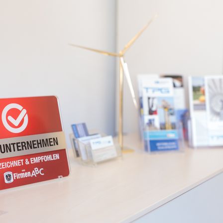 Ein rotes Schild mit einem Häkchen darauf steht 'Geprüftes Unternehmen von FirmenABC' auf einem weißen Schreibtisch mit Broschüren und einer Windmühle im Hintergrund.