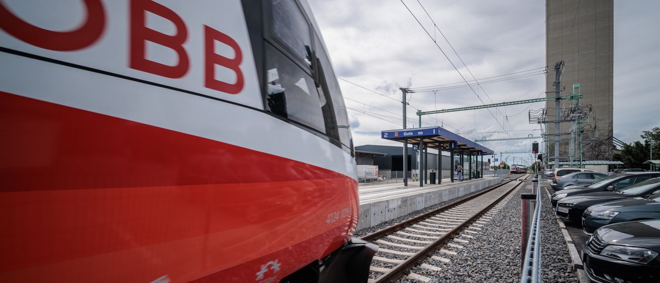 Ein BB-Zug nähert sich einem Bahnhof unter einem bewölkten Himmel. Die Vorderseite des Zuges ist in Rot und Weiß gehalten. Der Bahnhof hat ein blaues Vordach und ein Schild mit der Aufschrift 'Golis'.