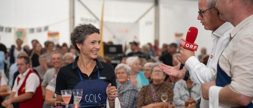 Eine ältere Frau in einer blauen Schürze mit dem Wort Gois darauf lächelt und hält einen Tablett mit Weinflaschen und Gläsern.