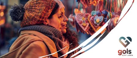 Two women wearing winter hats and scarves are standing close together, looking at colorful heart-shaped ornaments hanging from strings. The image has a white border and text reading 'THOMASMA' and 'Hauptplatz / Markt'.