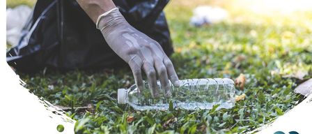 Eine Person mit Handschuhen hebt eine Plastikflasche vom Gras auf.