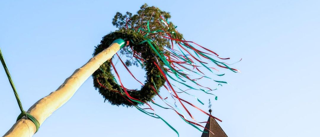 Ein großer Baumast mit einem Kranz, farbigen Bändern und einem Holzstab schwebt in der Luft über einer kleinen Kirche.