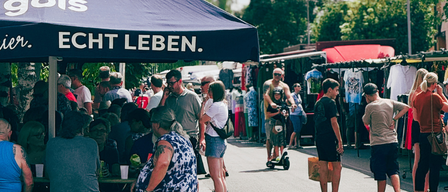 Ein Markt unter einem Regenschirm mit Leuten, die in Ständen stöbern, einem Mann mit einem Kind auf einem Roller und anderen, die an einem sonnigen Tag vorbeigehen.