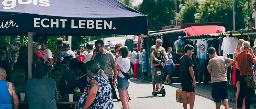 Ein Markt unter einem Regenschirm mit Leuten, die in Ständen stöbern, einem Mann mit einem Kind auf einem Roller und anderen, die an einem sonnigen Tag vorbeigehen.