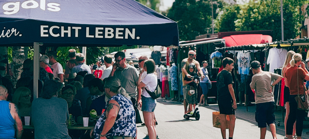Ein Markt unter einem Regenschirm mit Leuten, die in Ständen stöbern, einem Mann mit einem Kind auf einem Roller und anderen, die an einem sonnigen Tag vorbeigehen.