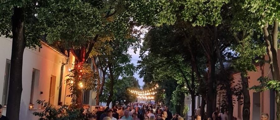 Eine von Bäumen gesäumte Straße ist mit Menschen gefüllt, die wahrscheinlich an einer Abendveranstaltung teilnehmen. Viele tragen Brillen und einige haben Getränke. Bäume sind mit Lichterketten beleuchtet.