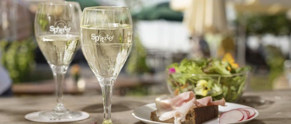 Zwei mit Wein gefüllte Weingläser stehen auf einem Tisch, begleitet von einem Teller mit Schinken und einer Schüssel Salat.