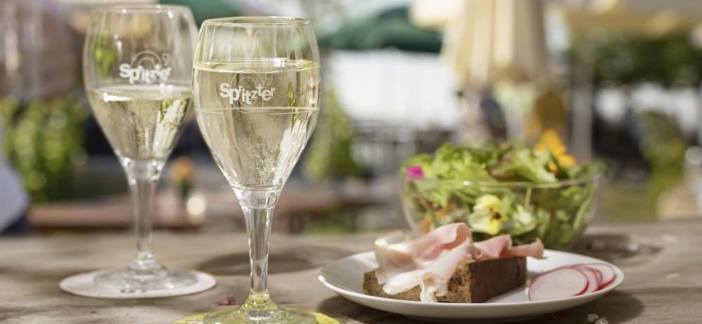 Zwei Weingläser mit 'Spitzer'-Etikett, gefüllt mit Weißwein, auf einem Tisch mit einer Platte mit Schinken und einem Salat.