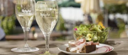 Two wine glasses with 'Spitzer' label, filled with white wine, on a table with a plate of ham and a salad.