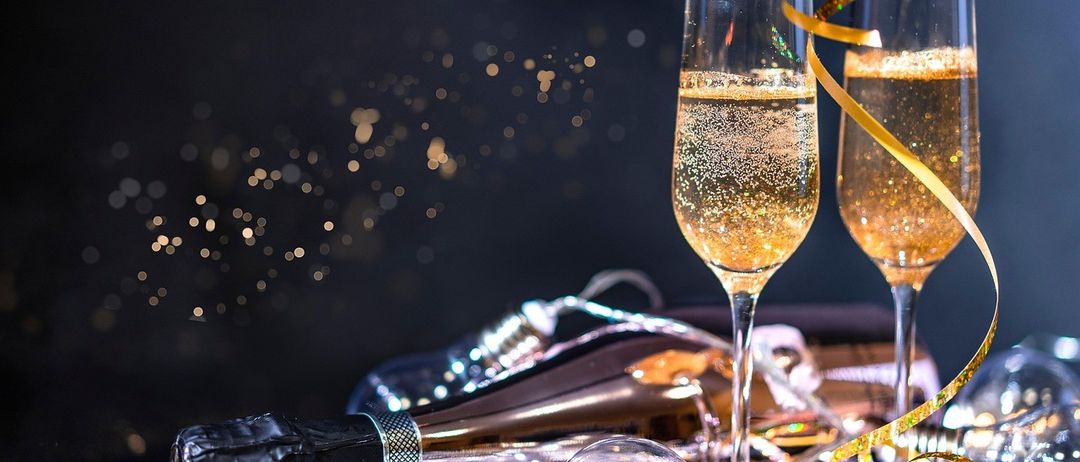 Zwei Gläser Champagner mit goldenen Blasen im Vordergrund. Eine Champagnerflasche und festliche Dekorationen sind in der Nähe. Der Hintergrund ist dunkel mit funkelnden Lichtern.