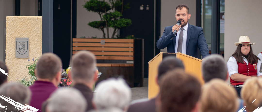 Ein Mann spricht in ein Mikrofon an einem Rednerpult vor einem Publikum. Dahinter steht ein Baum und eine Holzbank.