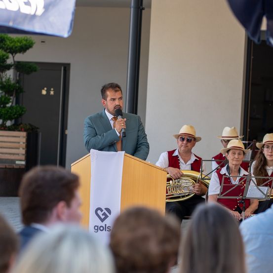Ein Mann spricht an einem Rednerpult mit Mikrofon, mit einer Band in traditioneller Kleidung hinter ihm. Das Publikum sitzt vor dem Rednerpult und beobachtet den Mann.
