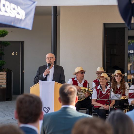Ein Mann spricht an einem Podium mit Mikrofon. Dahinter spielt eine Band von Musikern Instrumente. Ein Publikum sieht vorne zu. Eine Fahne und ein Gebäude mit einer Glastür sind im Hintergrund.