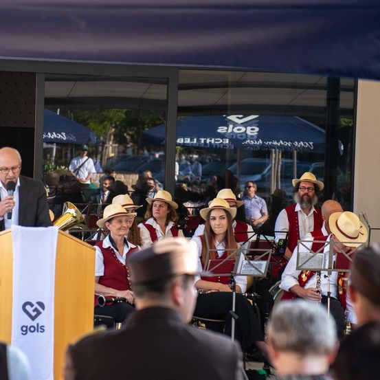 Eine Band spielt vor einer Menge, mit einem Mann am Podium. Die Musiker tragen Hüte und Westen. Das Publikum sitzt und steht. Im Hintergrund spiegelt eine Glaswand einen Schirm und Autos.