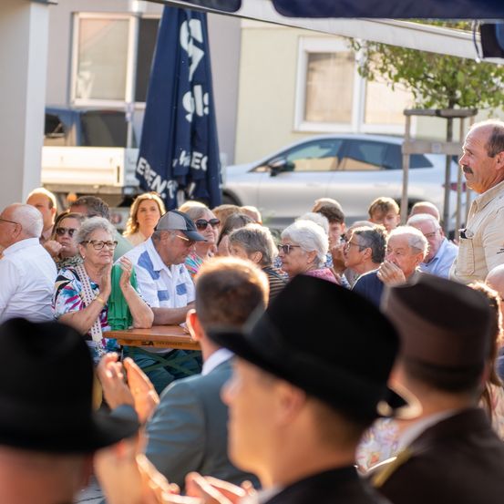 Eine Menschenmenge sitzt und steht draußen, einige tragen Hüte. Sie scheinen eine Veranstaltung zu beobachten, ein Mann lächelt und klatscht. Hinter ihnen steht ein blauer Schirm und ein geparktes Auto.