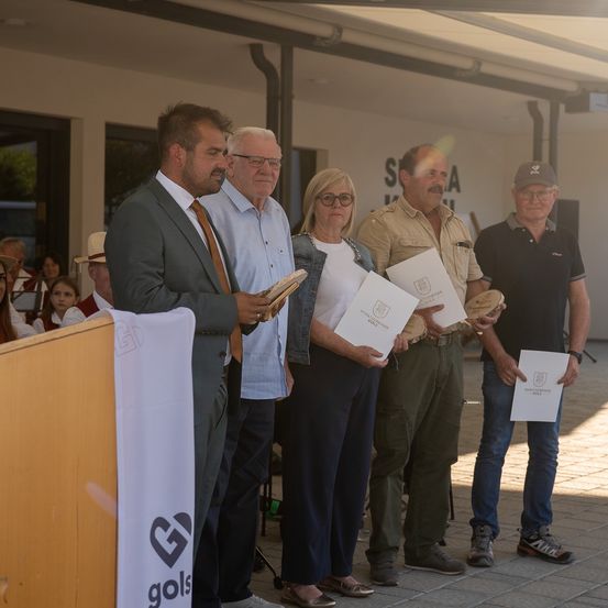 Fünf Personen stehen in einer Reihe, zwei halten Urkunden, eine weitere ein Buch, andere Trommeln, bei einer Veranstaltung im Freien.