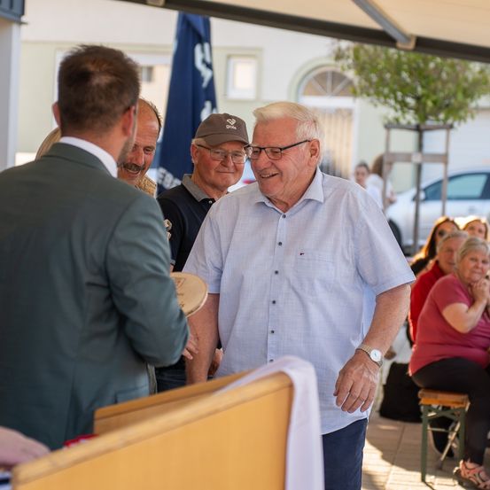 Eine Gruppe von Menschen steht draußen, ein älterer Mann mit Brille lächelt. In der Nähe spricht ein Mann im Anzug mit einer anderen Person. Ein Flagge ist im Hintergrund.