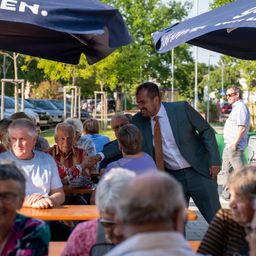 Ein Mann in einem Anzug begrüßt ältere Menschen, die unter Schirmen bei einer Veranstaltung im Freien sitzen. Autos und Bäume sind im Hintergrund zu sehen.