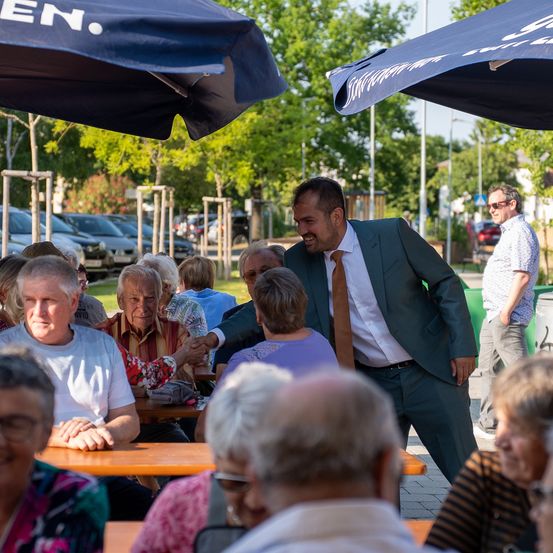 Ein Mann in einem Anzug begrüßt ältere Menschen, die unter Schirmen bei einer Veranstaltung im Freien sitzen. Autos und Bäume sind im Hintergrund zu sehen.