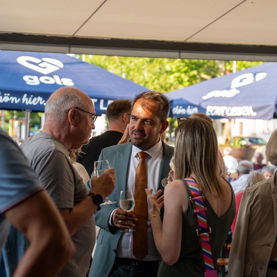 Mehrere Personen versammeln sich bei einer Veranstaltung im Freien. Ein Mann im Anzug hält zwei Gläser Wein, und eine Frau neben ihm trägt einen bunten Riemen. Im Hintergrund befinden sich blaue Zelte mit weißen Logos.